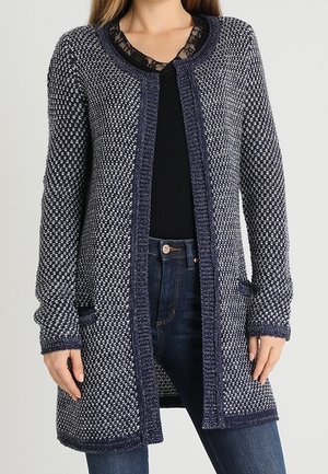 Vrouw die een lange marineblauwe en witte gebreide cardigan draagt over een zwart topje en donkerblauwe spijkerbroek, staand met handen ontspannen langs haar zij.
