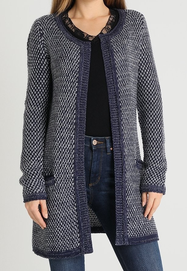 Femme portant un long cardigan en maille bleu marine et blanc sur un top noir et un jean bleu foncé, debout avec les mains détendues le long du corps.