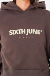 Sudadera gris oscuro con capucha. Presenta el texto "SIXTH JUNE" en crema y "PARIS" debajo en una fuente más pequeña. Textura de tela suave.