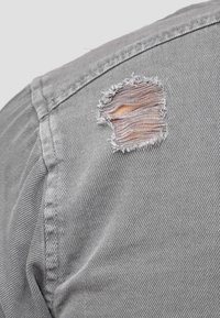 Grijs stoffen overhemd met zichtbare stiksels en een beschadigde scheur op de schouder, waarbij een lichtere binnenlaag zichtbaar is. Over het geheel gladde textuur.