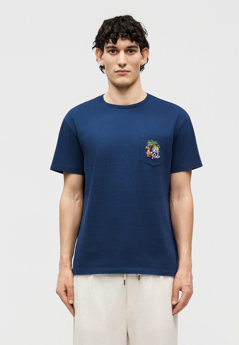 Άνδρας που φορά μπλούζα σε σκούρο μπλε χρώμα με πολύχρωμο τροπικό σχέδιο και τη λέξη «Polo» στην τσέπη στο στήθος, συνδυασμένη με λευκό παντελόνι.