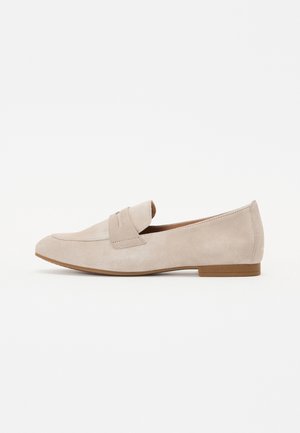 Beige mokkasins i ruskind med lav hæl og remdetalje over toppen, vist i profil mod hvid baggrund.