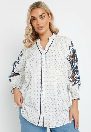Weiße Bluse mit maritimen Punkten, bestickten Blumendetails an den Ärmeln, Knopfverschluss vorne und Rüschendetails am Kragen. Lockere Passform.