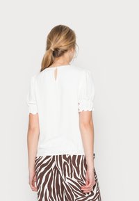 Blouse blanche à manches bouffantes avec un détail dos en forme de trou de serrure. Tissu lisse avec une coupe ample, associée à un pantalon rayé marron et blanc.