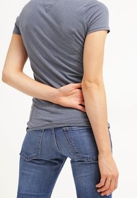 Camiseta ajustada gris de manga corta, combinada con pantalones vaqueros azules. La camiseta tiene una textura sutil y un detalle de escote en V en la parte de atrás.
