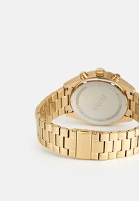 Montre en acier inoxydable doré avec un bracelet à maillons, un cadran rond et des fonctionnalités chronographe. Comprend un fond de boîtier transparent affichant le logo de la marque.