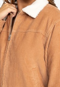 Veste en velours côtelé orange-brun avec un col en polaire blanche. Dotée d'une fermeture éclair, d'une texture côtelée verticale et d'une poche latérale.