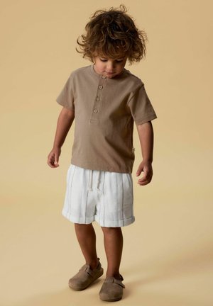 Jeune enfant aux cheveux bouclés portant une chemise marron boutonnée, un short blanc rayé et des chaussures marron à enfiler, debout devant un fond beige.