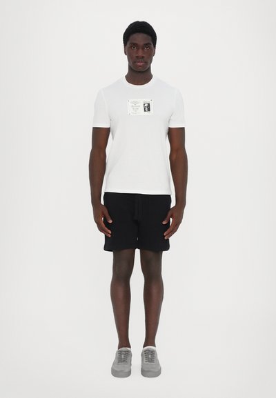 Tricou alb cu mânecă scurtă și imprimeu grafic, asortat cu șorturi negre și adidași gri. Modelul stă în picioare pe un fundal simplu.