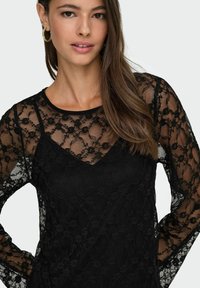Czarna koronka z kwiatowym wzorem, przejrzystymi długimi rękawami i dekoltem w kształcie litery V. Materiał jest teksturowany, lekki i elegancki.