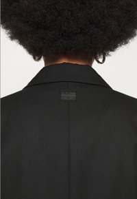Personne aux cheveux bouclés portant une veste noire avec une étiquette G-Star RAW sur le haut du dos et des boucles d'oreilles dorées en forme d'anneau, vue de dos.
