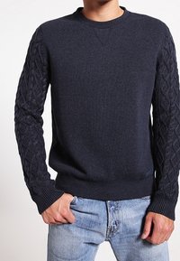 Marinblå sweatshirt med kabelstickat mönster på ärmarna, ribbade muddar och nederkant, rund hals, mjuk textur, bärs med ljusblå jeans.