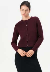 Donna che indossa un cardigan buttoned di un viola scuro e una gonna nera, in piedi con le mani incrociate e guardando in basso a sinistra.