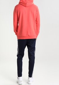 Felpa color corallo con cappuccio, abbinata a pantaloni della tuta blu navy con strisce bianche sui lati; entrambi in un misto cotone, che mostrano una vestibilità rilassata.