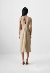 Femme aux longs cheveux foncés portant une robe midi beige sans manches avec un dos en V profond et des sandales à talons, debout devant un fond clair uni.