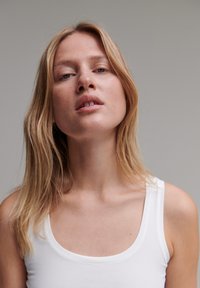 Femme blonde aux cheveux lâchés, portant un débardeur blanc, regardant légèrement vers le haut sur un fond gris uni.