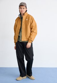 Giacca marrone chiaro con polsini elasticizzati, indossata sopra una camicia verde, abbinata a pantaloni neri e scarpe beige. Cappellino da baseball grigio aggiuntivo.