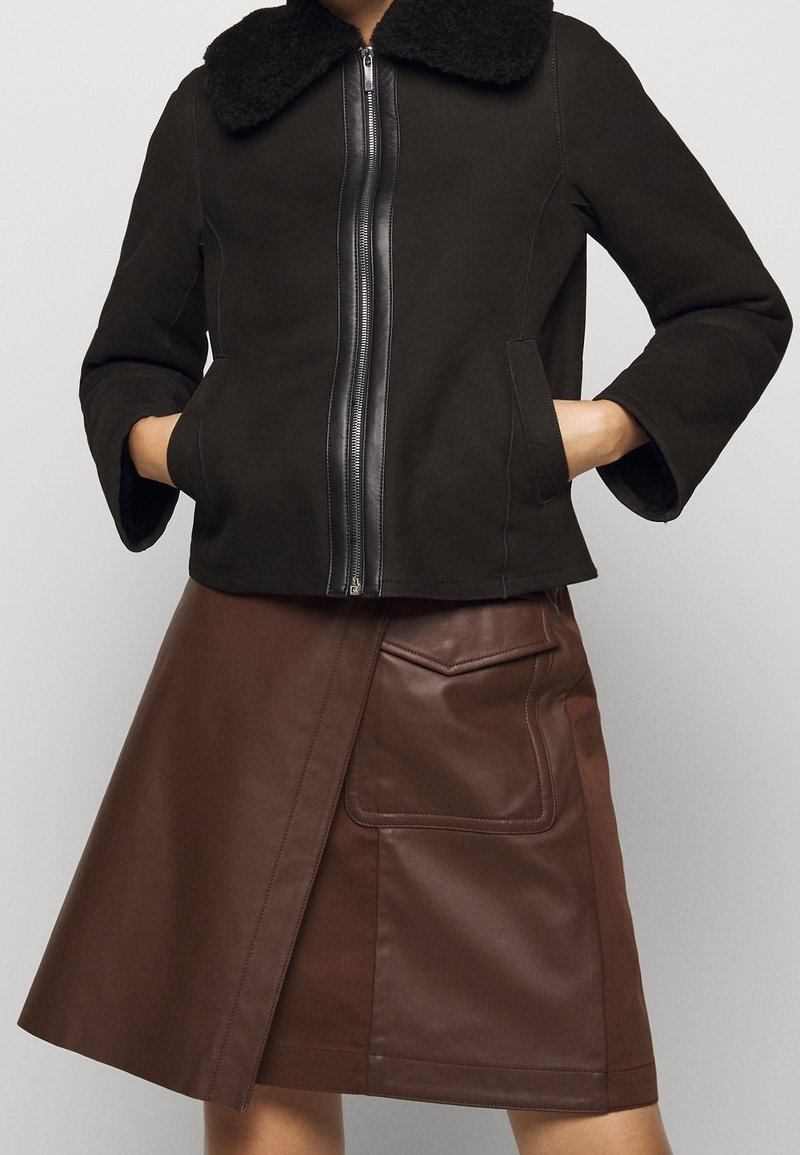 Femme portant une veste noire zippée avec un col duveteux et une jupe en cuir marron avec une grande poche à l’avant. Mains dans les poches de la veste.