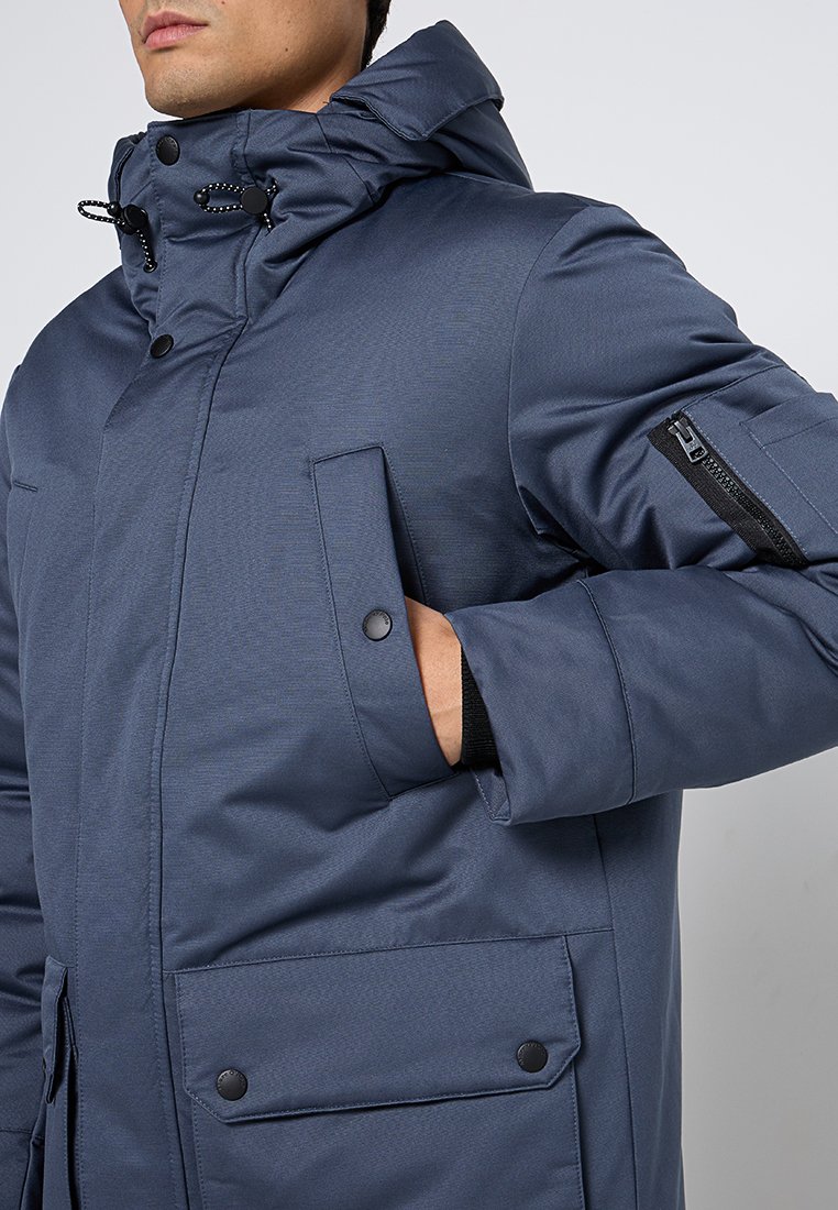 Marineblauwe gewatteerde jas met een hoge kraag, verstelkoord, voorzakken en een ritszak op de mouw met zwarte accenten.