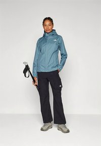 Šviesiai mėlyna neperšlampama striukė su gobtuvu, priekyje su užtrauktuku ir logotipu ant krūtinės, derinama su juodomis kargo kelnėmis ir pilkais žygių batais.