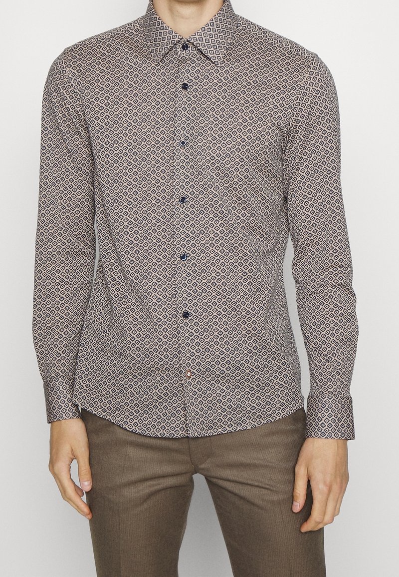 Homme portant une chemise ajustée à manches longues avec un petit motif géométrique en losanges et un pantalon marron.