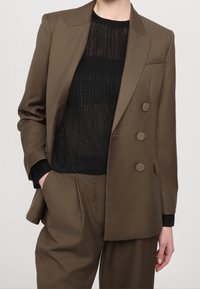 Person som bär en brun dubbelknäppt kavaj och matchande byxor med en svart halvgenomskinlig räfflad stickad topp, hand i fickan.