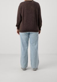 Felpa marrone, strutturata, con una vestibilità rilassata e polsini a coste, abbinata a jeans in denim blu chiaro a gamba larga. Sneakers grigio chiaro visibili.