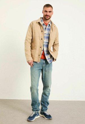 Beige Baumwolljacke über einem karierten Hemd, orangen T-Shirt, hellblauen Jeans und marineblauen Turnschuhen. Minimalistisches Design, lässige Passform.