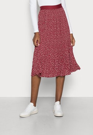 Femme portant une jupe midi rouge à fleurs avec une large ceinture, associée à des baskets blanches et un haut blanc à manches longues.