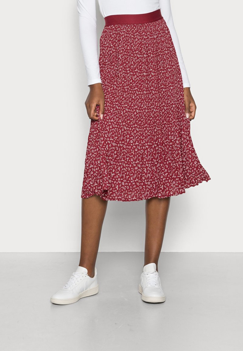 Femme portant une jupe midi rouge à fleurs avec une large ceinture, associée à des baskets blanches et un haut blanc à manches longues.