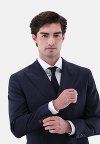 Junger Mann, der die Manschette seines dunkelblauen Anzugs justiert, trägt ein weißes Hemd und eine schwarze Krawatte, mit ordentlich gestyltem dunklem Haar vor einfarbigem Hintergrund.