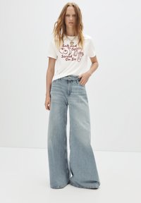 Witte t-shirt met rode grafische tekst en afbeeldingen. Lichtblauwe wijde jeans met hoge taille en riem. Glad denimtextuur, casual pasvorm.