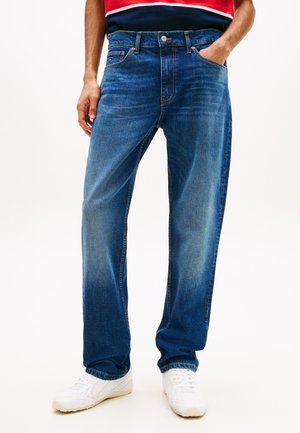 Mand iført afslappede blå jeans og hvide sneakers, stående med den ene hånd i lommen mod en ensfarvet baggrund.