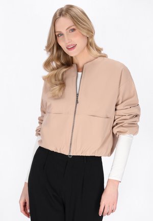 Vrouw met blond golvend haar draagt een beige cropped jas met rits over een wit shirt met lange mouwen en zwarte broek met hoge taille, glimlachend tegen een witte achtergrond.