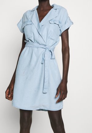 Robe en denim bleu clair à manches courtes avec un design croisé, deux poches poitrine et une ceinture à nouer à la taille. Texture lisse, coupe décontractée.