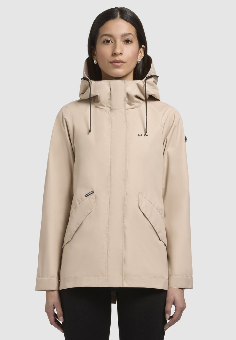 Beige waterdichte jas met een hoge kraag en verstelbare capuchon, voorzien van twee grote voorzakken en een gladde, matte textuur.