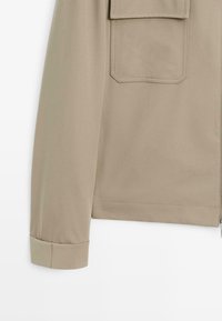Beige jas van een gladde, lichte stof, met een enkele patchzak op de linkermouw en een oprolbare manchettenontwerp.