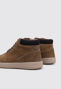 Zapatos altos de ante marrón con un collar acolchado negro, diseño de cordones y suela de goma beige. Presentan un detalle de logo en el lateral.