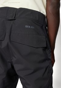 Sorte vanntette bukser med en glatt tekstur, med baklommer og en "RAIN.RDY" logo i lys blå. Justerbar midje kan sees.