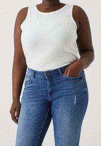 Camiseta sin mangas blanca de canalé combinada con jeans azules de talle alto. La camiseta tiene un cuello redondeado y los jeans presentan un diseño clásico de cinco bolsillos.