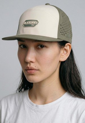 Young person with long dark hair wearing a beige and olive "BLACKSKIES PRO" cap and a plain white shirt, facing forward with neutral expression.