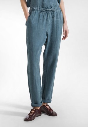 Osoba nosząca luźne niebieskie spodnie z elastycznym paskiem i troczkiem, ręce w kieszeniach, w połączeniu z ciemnoczerwonymi butami na sznurowadła.