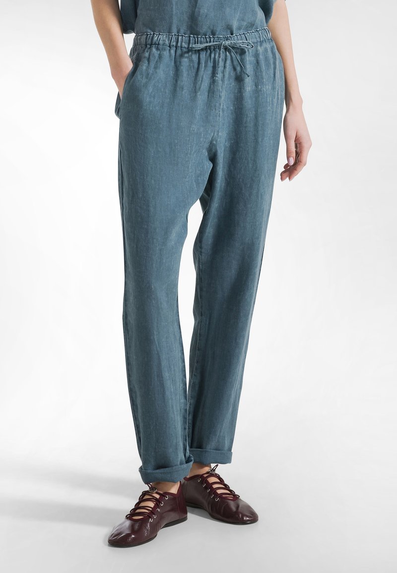 Persoon draagt blauwe, losvallende broek met elastische tailleband en koord, handen in zakken, gecombineerd met donkerrode veterschoenen.