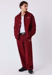 Completo giacca e pantaloni color bordeaux, con design a bottoni, tasche laterali e dettagli con logo, abbinato a sneakers nere su uno sfondo semplice.