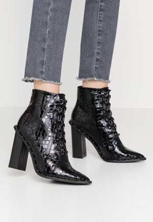 Une personne porte des bottines noires brillantes à lacets, à texture façon peau de crocodile, avec un bout pointu et un talon épais, assorties à un jean gris effiloché.