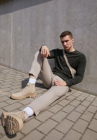 Stivaletti beige con tomaia testurizzata, pantaloni aderenti color beige chiaro e una maglietta a maniche lunghe verde scuro. Borsa a tracolla minimalista in beige.