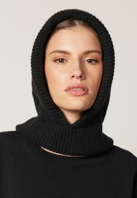 Svart stickad huvtröja med en ribbad design och hög krage. Materialet verkar mjukt och varmt, med en åtsittande form runt ansiktet.