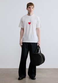 Λευκό T-shirt με κοντά μανίκια, διακοσμημένο με ένα γραφικό κόκκινης καρδιάς και την επιγραφή "HUGO" από κάτω, συνδυασμένο με σκούρο τζιν παντελόνι φαρδιάς γραμμής και στρογγυλή μαύρη τσάντα.