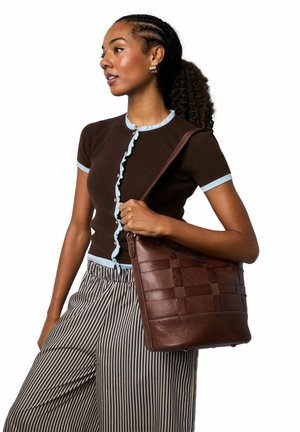 Bolso de cuero marrón con diseño trenzado, textura suave y base plana. La modelo lleva una blusa marrón acanalada con acentos azules y pantalones a rayas.