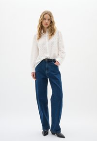 Camicia bianca con colletto, decorata con un motivo a occhio, abbinata a jeans blu dal taglio morbido. Il modello indossa una cintura nera e scarpe nere con punta.
